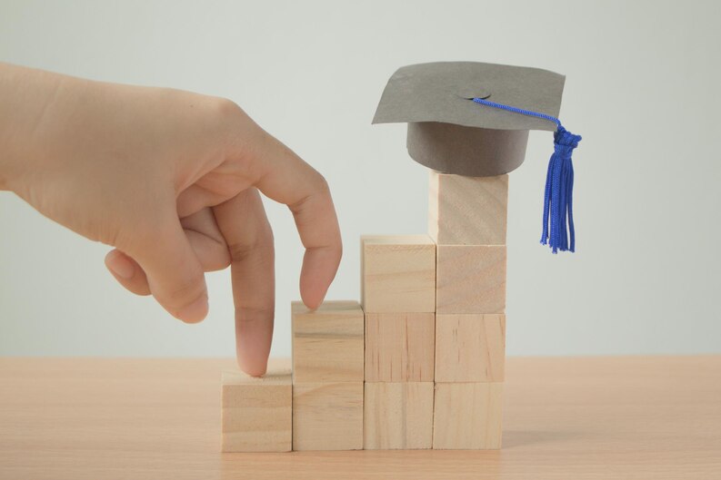 Hand walking up wooden block steps with a graduation cap on top, symbolizing career growth through education.